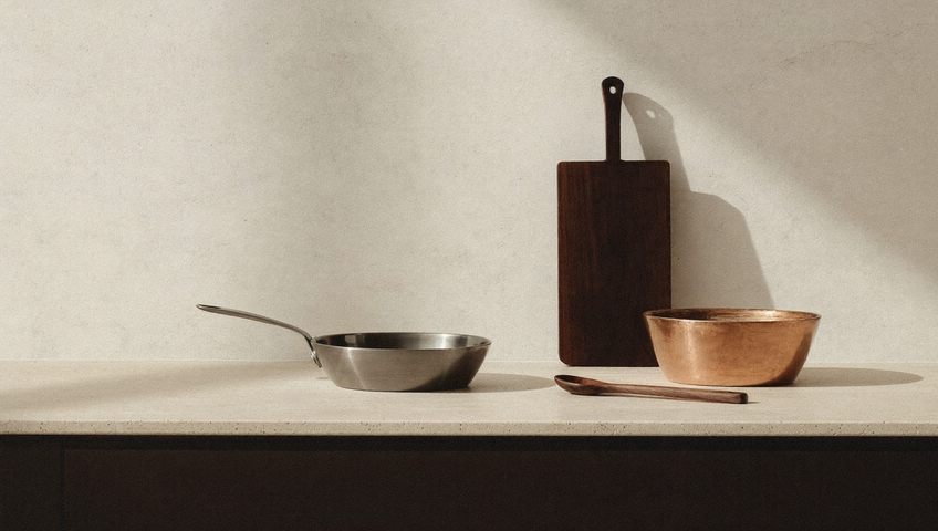 Su un banco di travertino beige, davanti a una parete in calce, una casseruola in acciaio spazzolato, una pentola in rame, un tagliere in noce appoggiato in verticale e un cucchiaio in legno d'ulivo in diagonale.