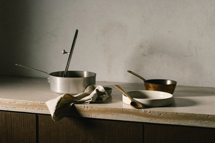 Luce di fine pomeriggio su un banco di travertino: pentola in acciaio con coperchio socchiuso, panno di lino piegato, cucchiaio in ulivo su un piatto in gres e una piccola casseruola in rame.